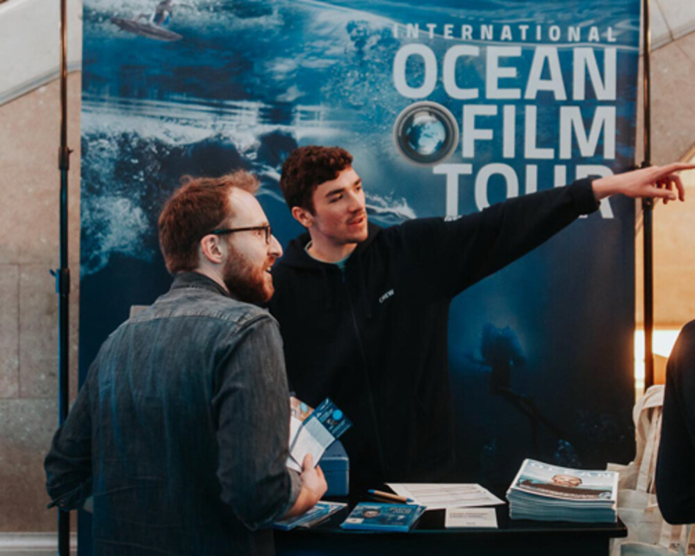 International Ocean Film Tour booth with staff providing information and handing out magazines to visitors.