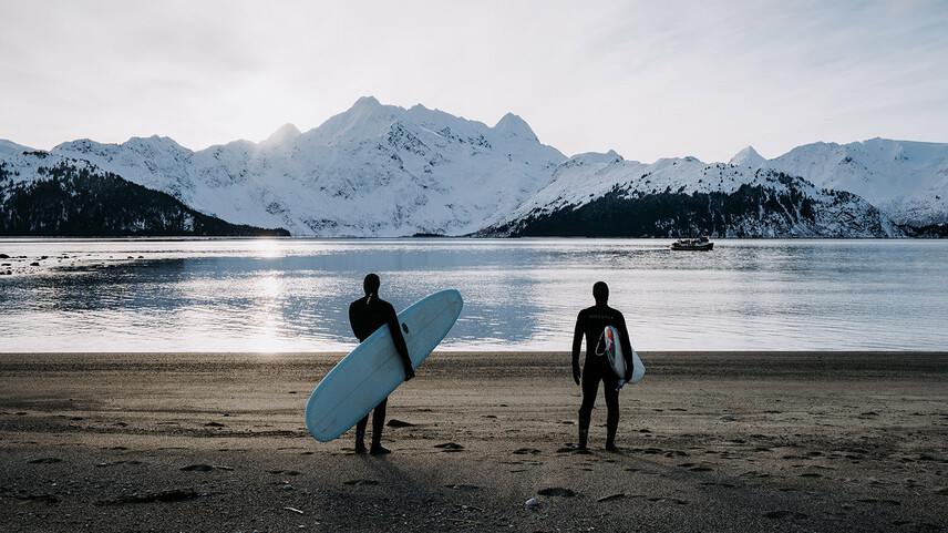 Two men in wetsuits and surfboards stand on a beach and look out over snow-covered mountains