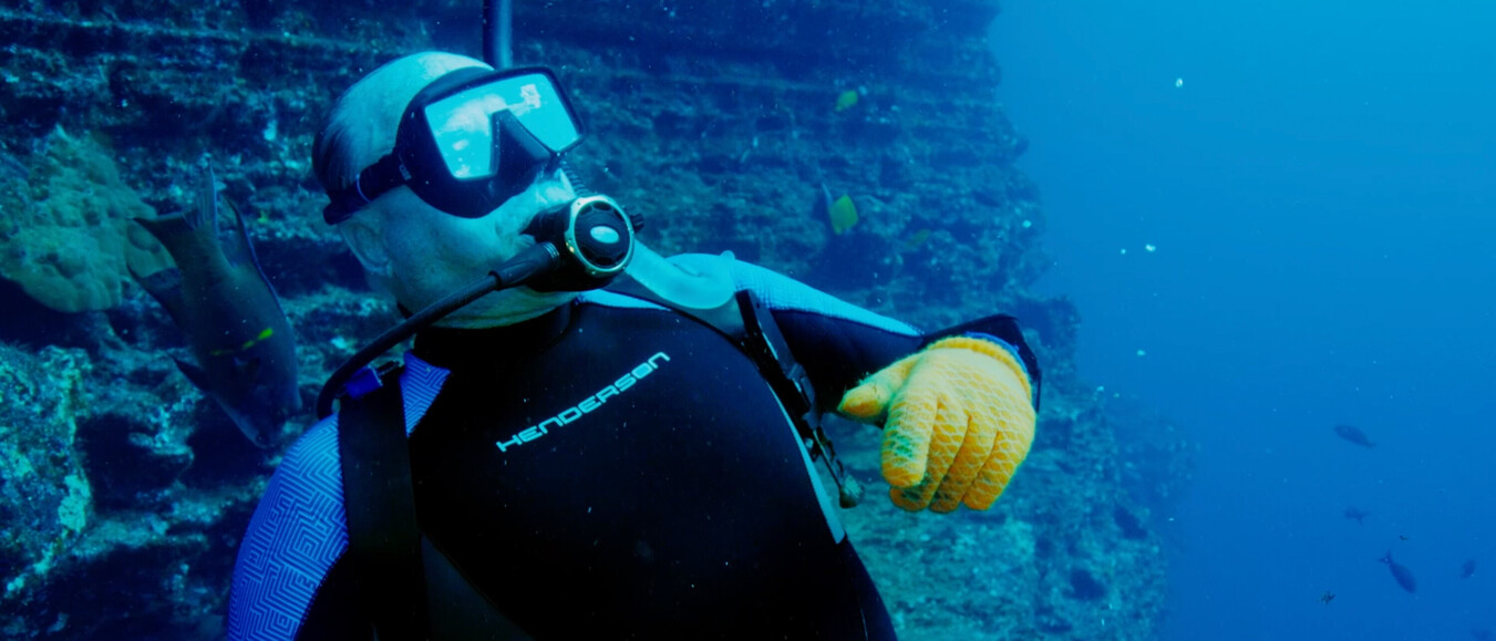 Older diver with yellow gloves and full scuba gear gesturing near an underwater reef